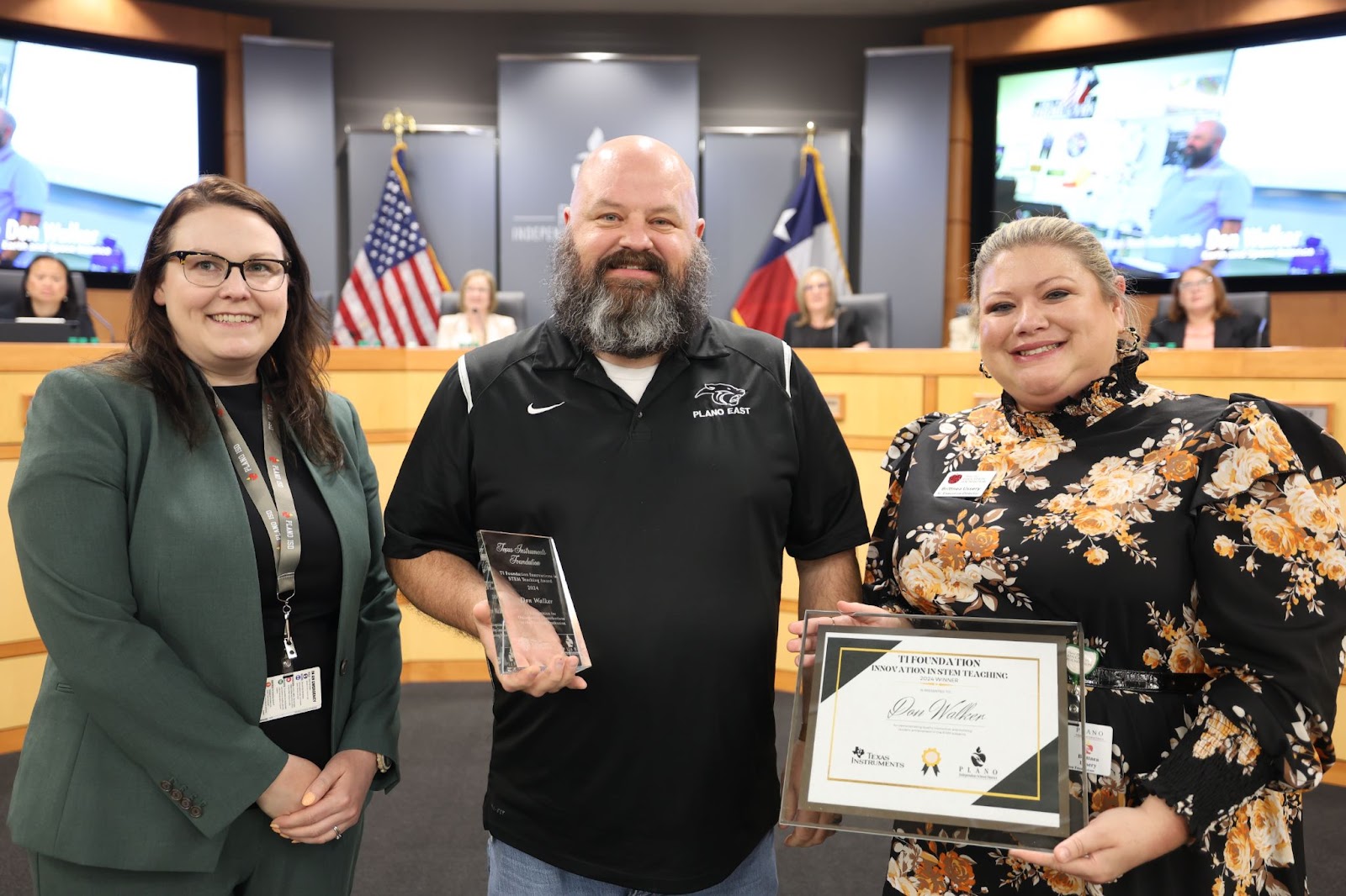 three individuals standing in front of the Plano ISD boardroom, the middle man is holding a certificate and plaque
